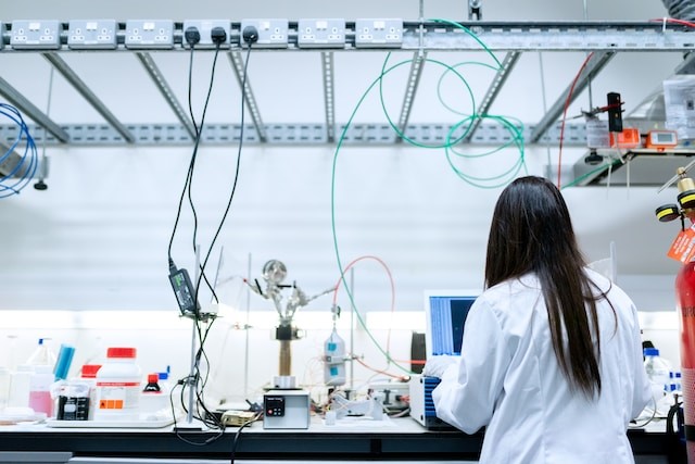 Een wit belicht lab met verschillende apparatuur. Er is een persoon met lang haar aan het werk achter een laptop.